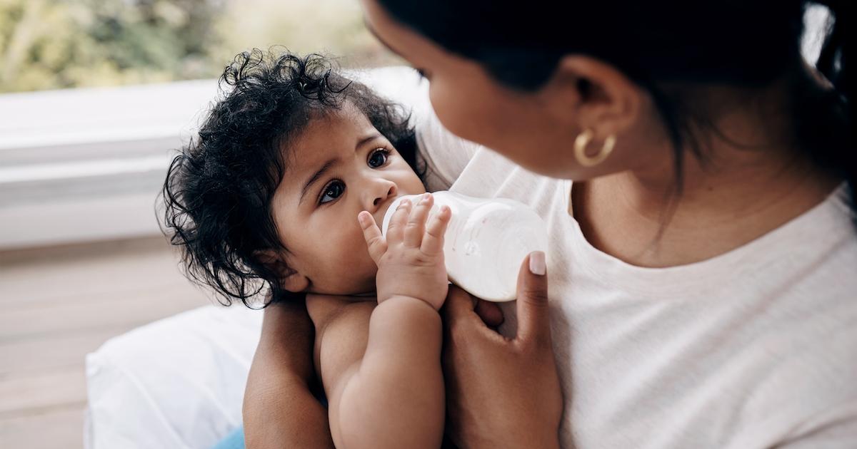 A baby drinks from a bottle and gazes up at their mother.