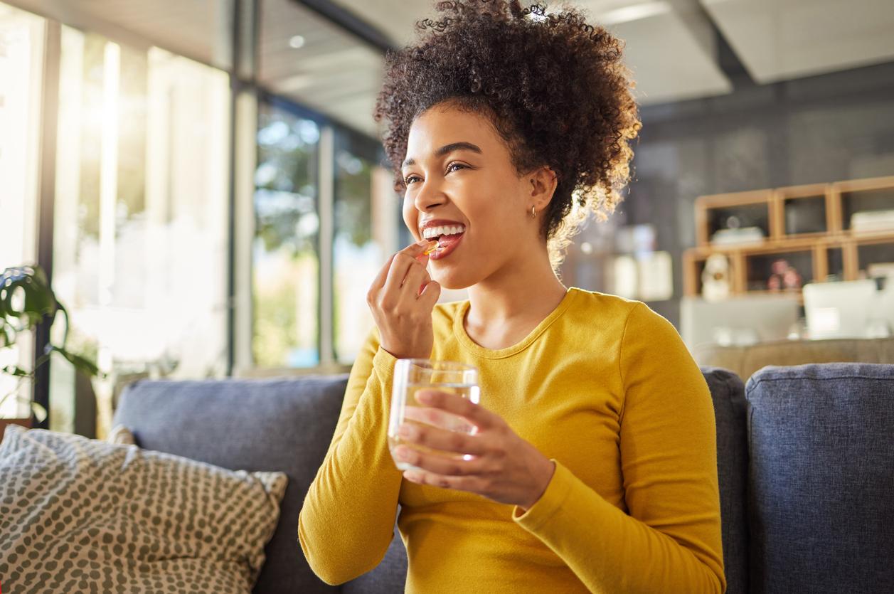 A woman in a yellow shirt holds a glass of water in one hand and brings a pill up to her open mouth with her other hand.