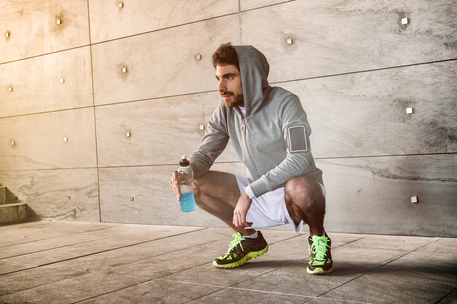 A man on a run crouches on the ground while holding a blue drink. 