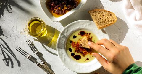 Photo of hand dipping bread into a white dish full of olive oil on a white surface
