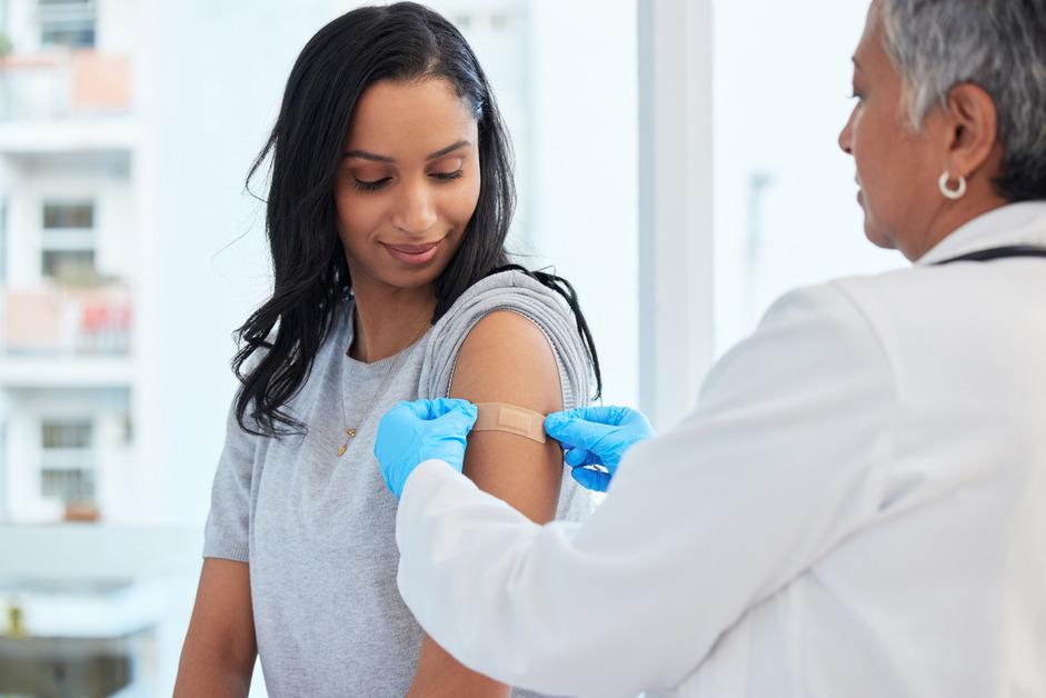 A woman with dark hair sits in the doctors office while a female doctor puts a bandage on her arm after receiving a vaccine. 