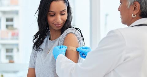 A woman with dark hair sits in the doctors office while a female doctor puts a bandage on her arm after receiving a vaccine.