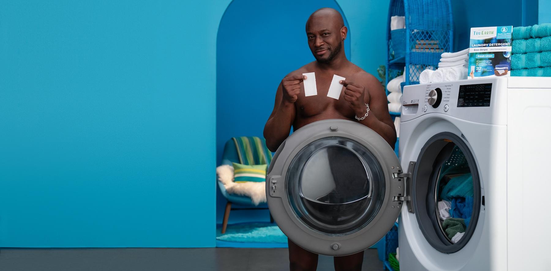 Taye Diggs topless, smirks and stands nude with a washing machine blocking his body, and holds up Tru Earth's laundry eco-strips