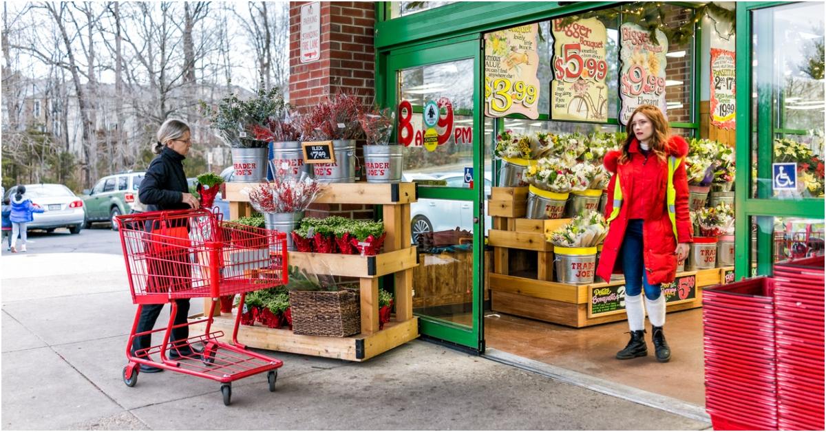 One woman with a red cart looks at plants outside a Trader Joe's while another woman in a red coat walks out of the store. 