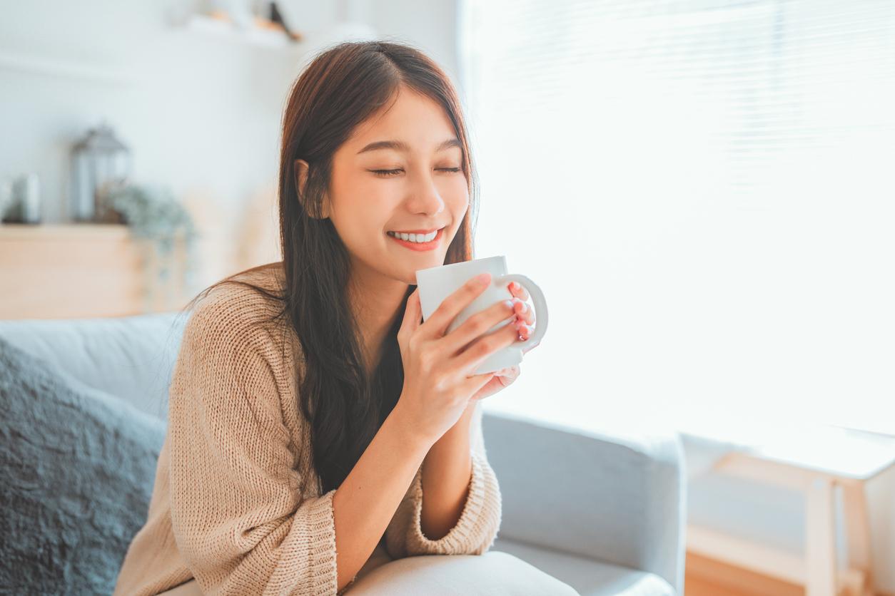 A smiling woman sips lemon gunpowder tea from a white mug on her bed.