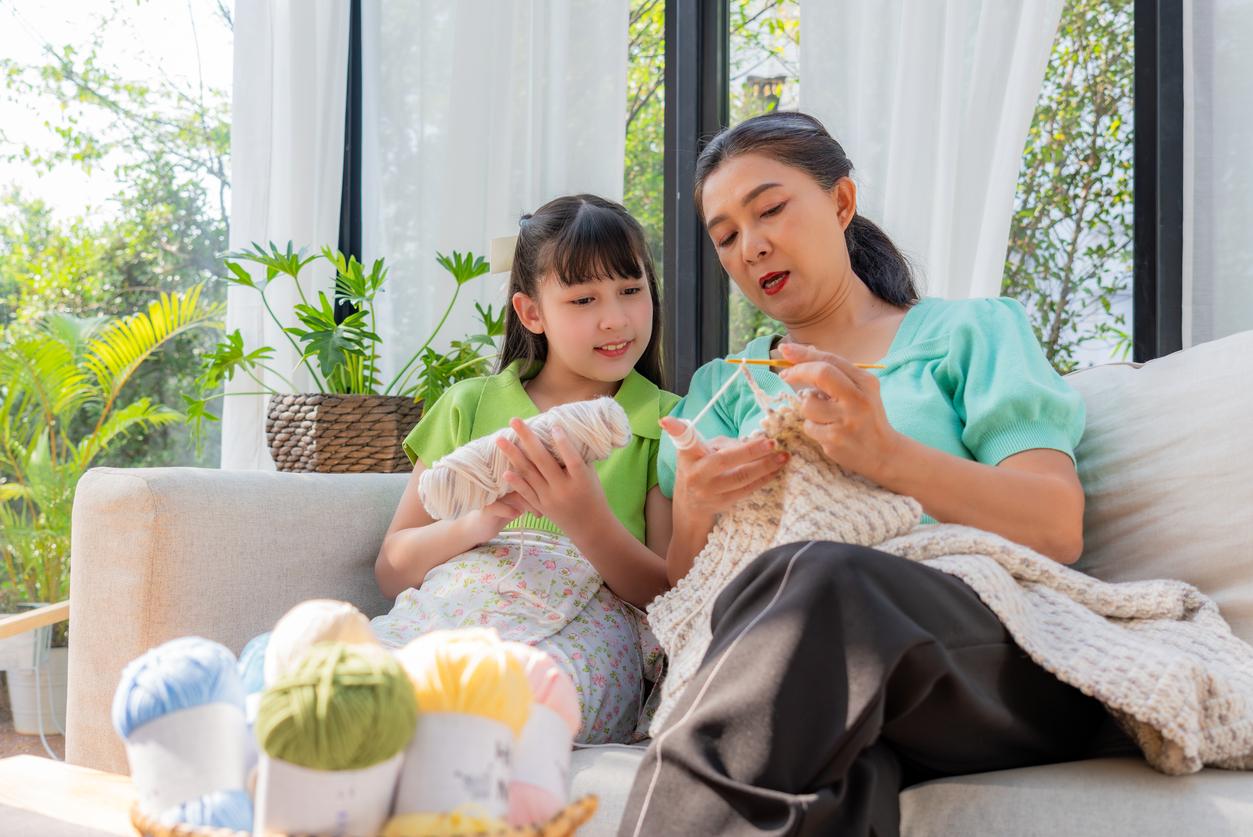 A young child and her family member crochet on the couch together beside plants and windows..