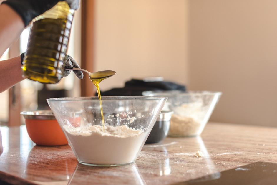 A person pouring a spoon of oil into a bowl of flower on a table with several other bowls on top. 