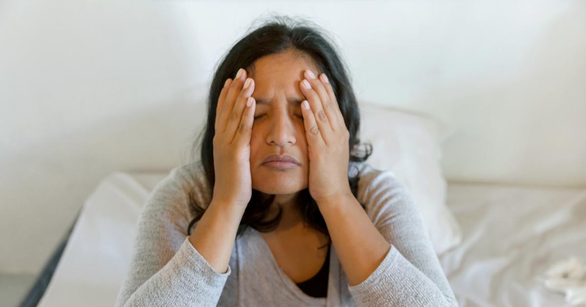 A woman wearing a gray shirt sits on a bed and grabs her face in a tired expression. 