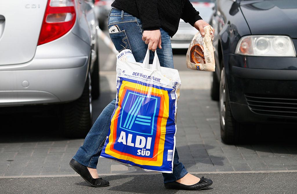 A shopper is pictured in a parking lot holding an Aldi grocery bag, with cars in the background.