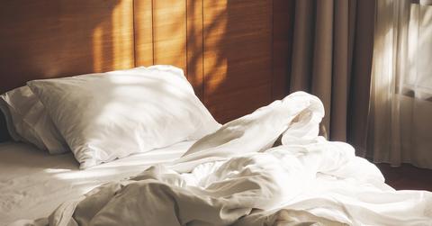An unmade bed with white sheets and a white duvet and pillow in the morning sunlight.