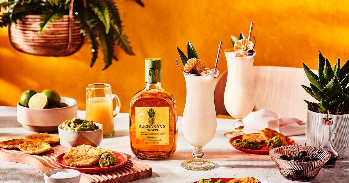 A bottle of Buchanan's Pineapple surrounded by cocktails and plates of food on a white table.