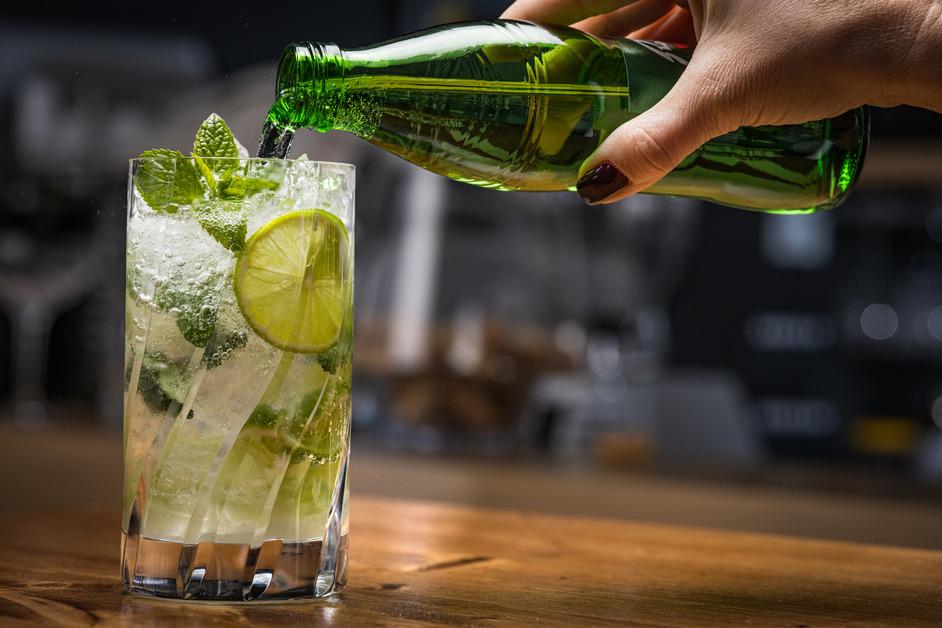 A close-up photo of someone pouring sparkling water into cocktail with limes and mint leaves. 