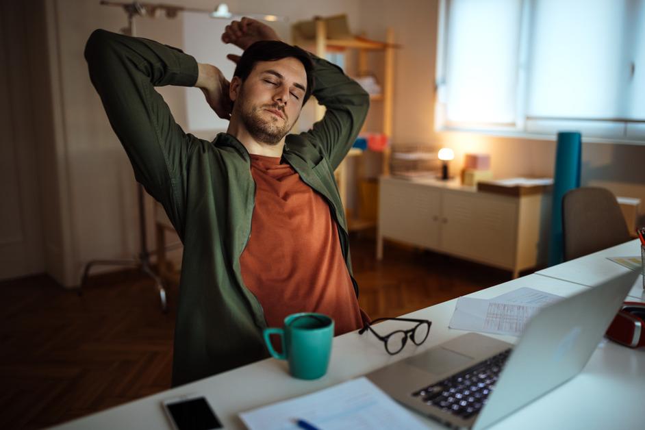 Man sits at a desk in a orange and green shirt while stretching his arms over his head. 