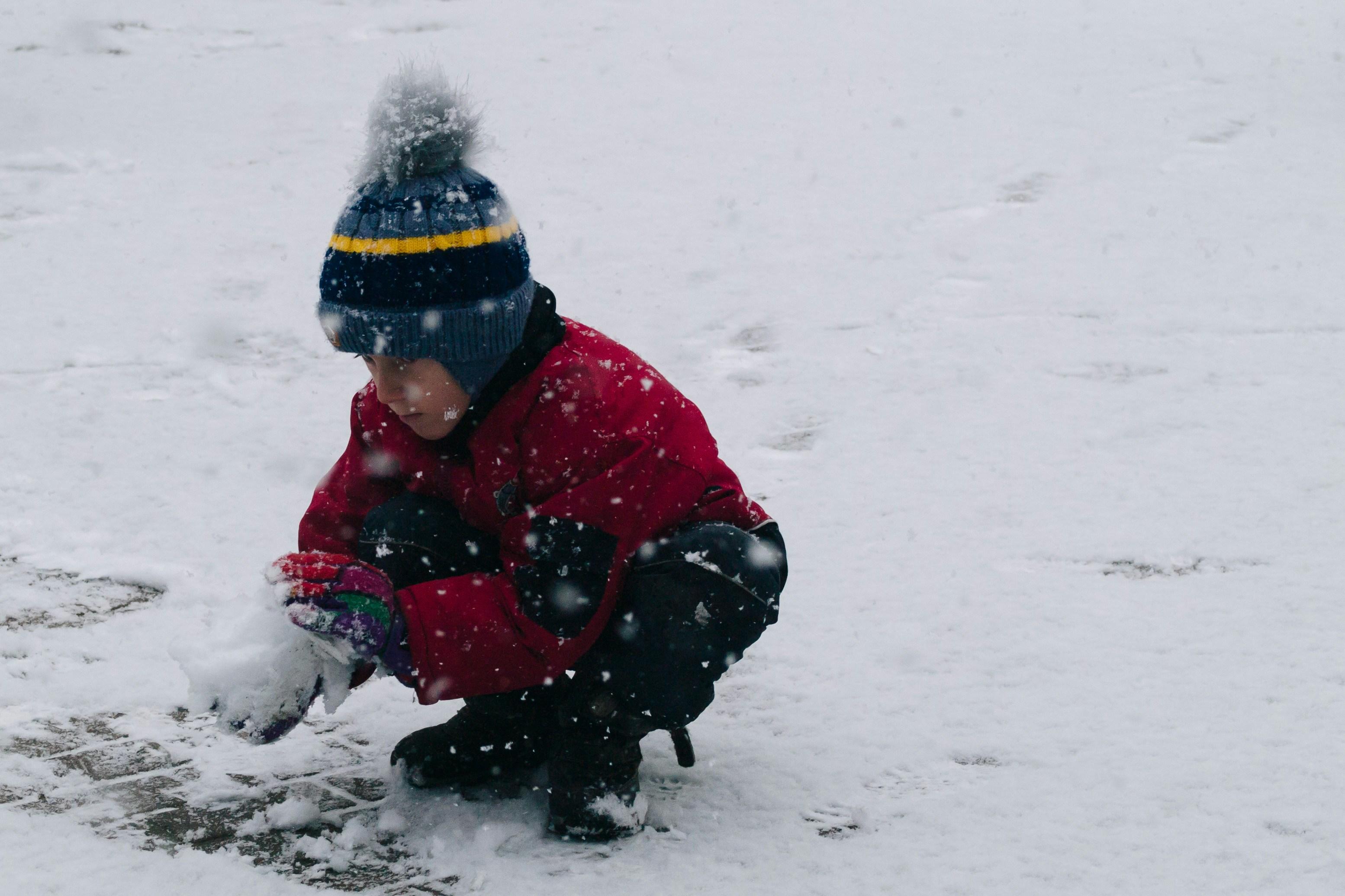 A young boy squats in the snow as he makes a snowball.