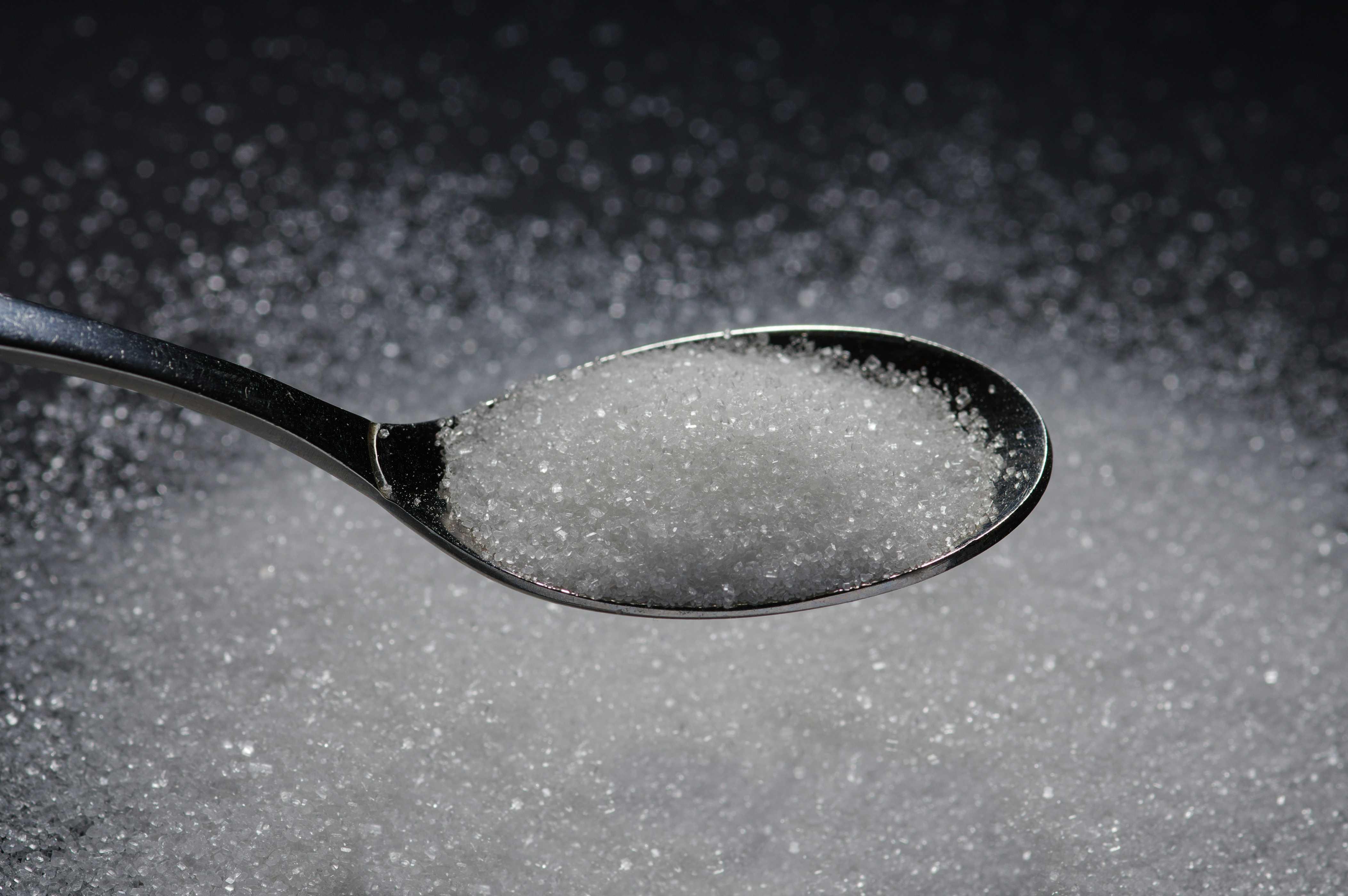 A spoonful of crystallized sugar is pictured above crystallized sugar on top of a table. 