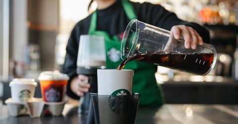 A Starbucks employee wearing a Starbucks uniform pours a black liquid into a personal white cup within a larger black container in a Starbucks store.
