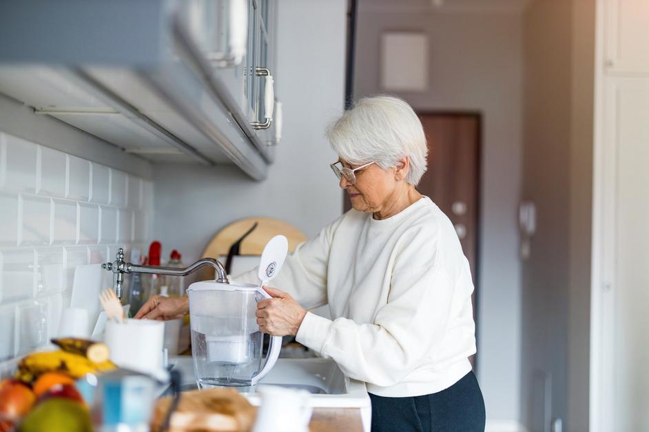 A photo of a senior woman in a kitchen filling a plastic water filter under the faucet. 