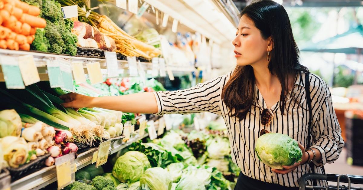 Woman in striped blouse holds a head of lettuce and reaches for a leek in a grocery store