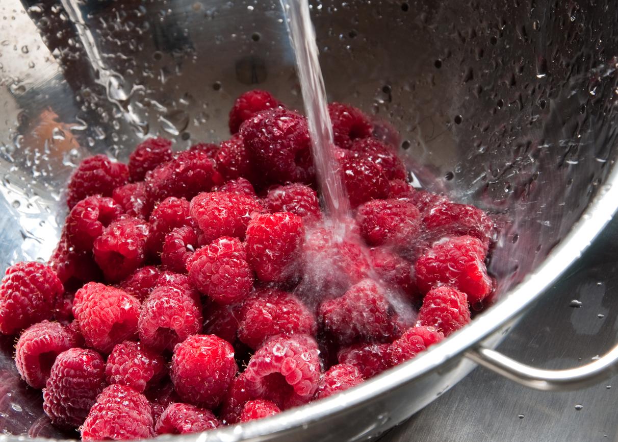 Raspberries in a strainer under a stream of water in the sink.