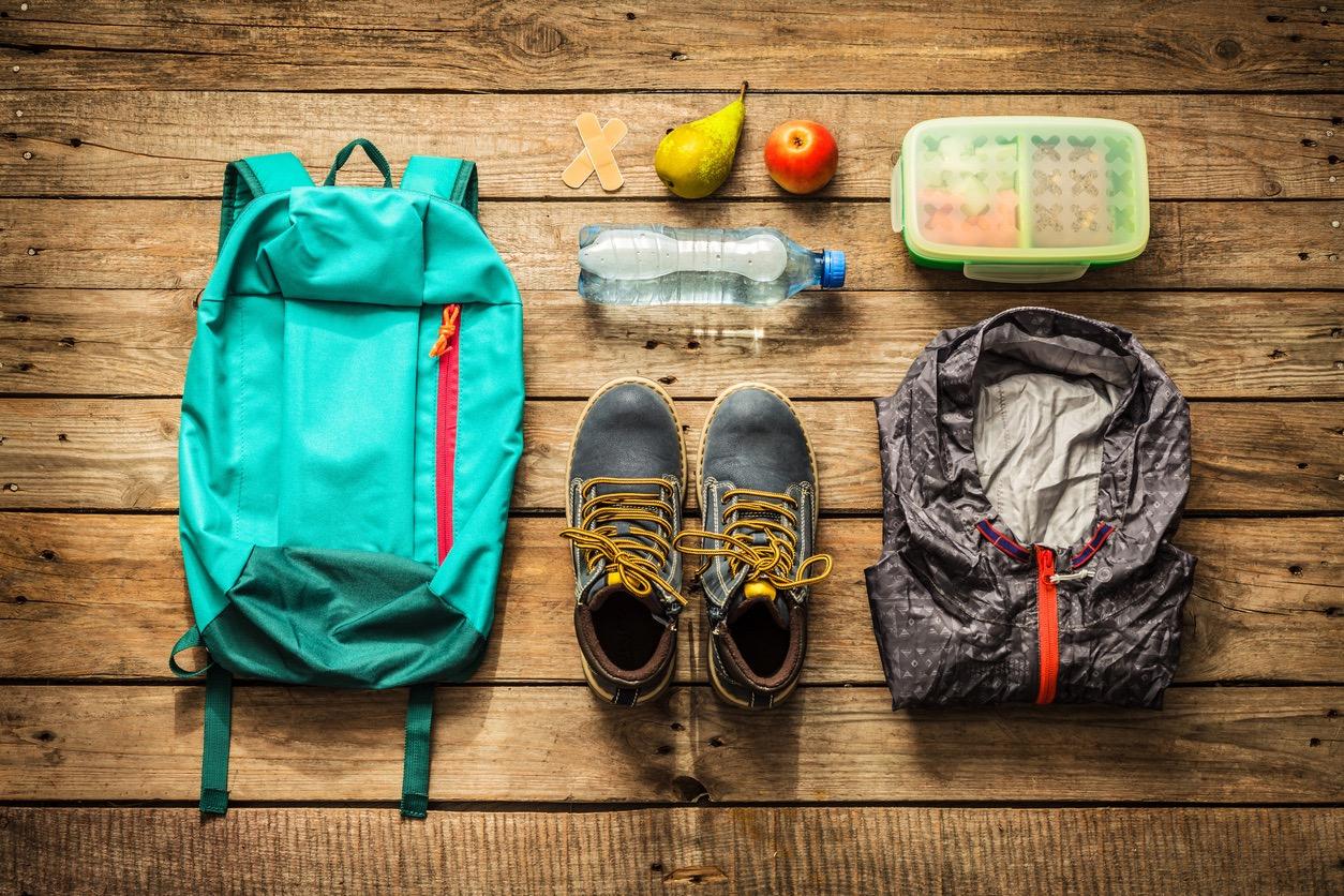 A blue backpack, hiking boots, raincoat, container with food, plastic water bottle, apple, pear, and bandaids laid out on a table.