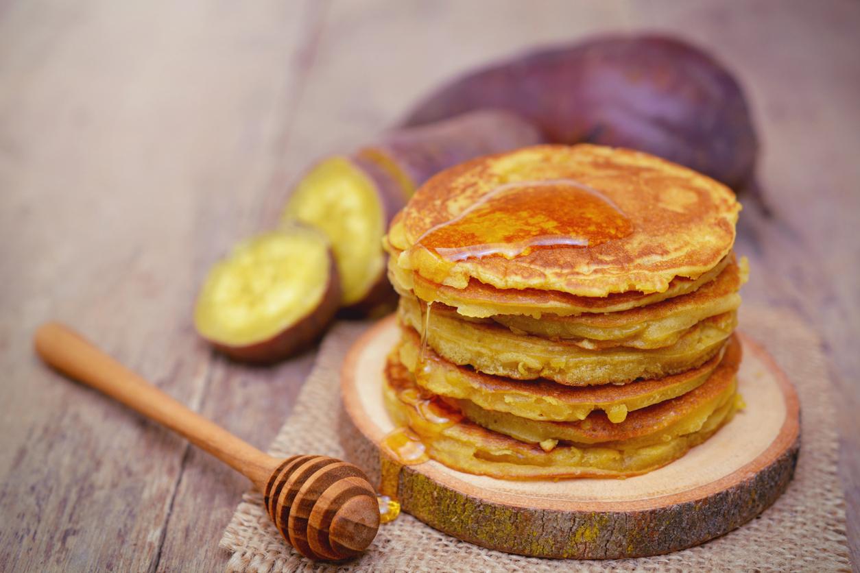 Sweet potato pancakes are pictured with a sliced sweet potato in the background and a wooden spoon with vegan honey on it.