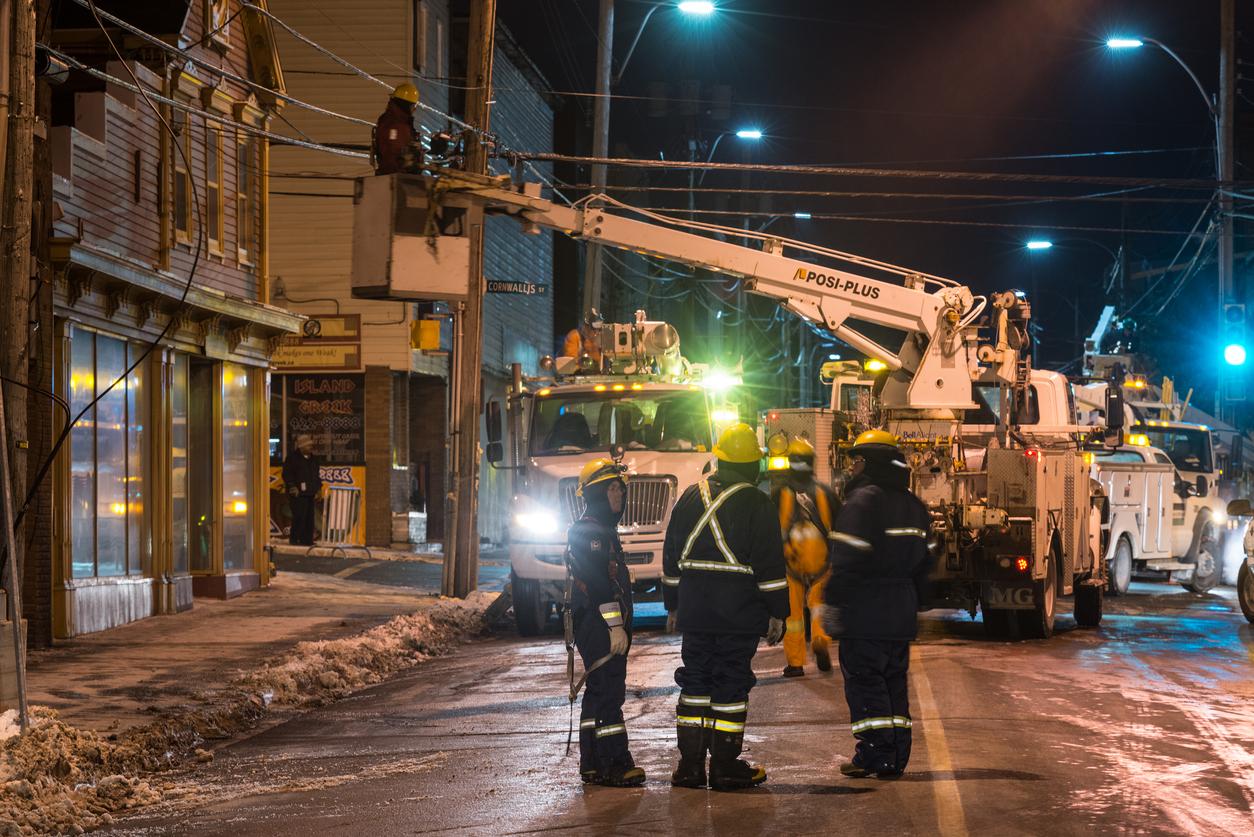 Three electricians standing next to a truck with a crane fixing a telephone pole.