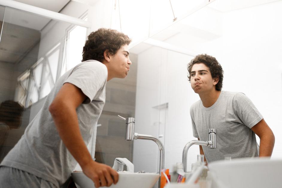 A man wearing gray stands in the bathroom while rinsing his mouth and looking in the mirror. 