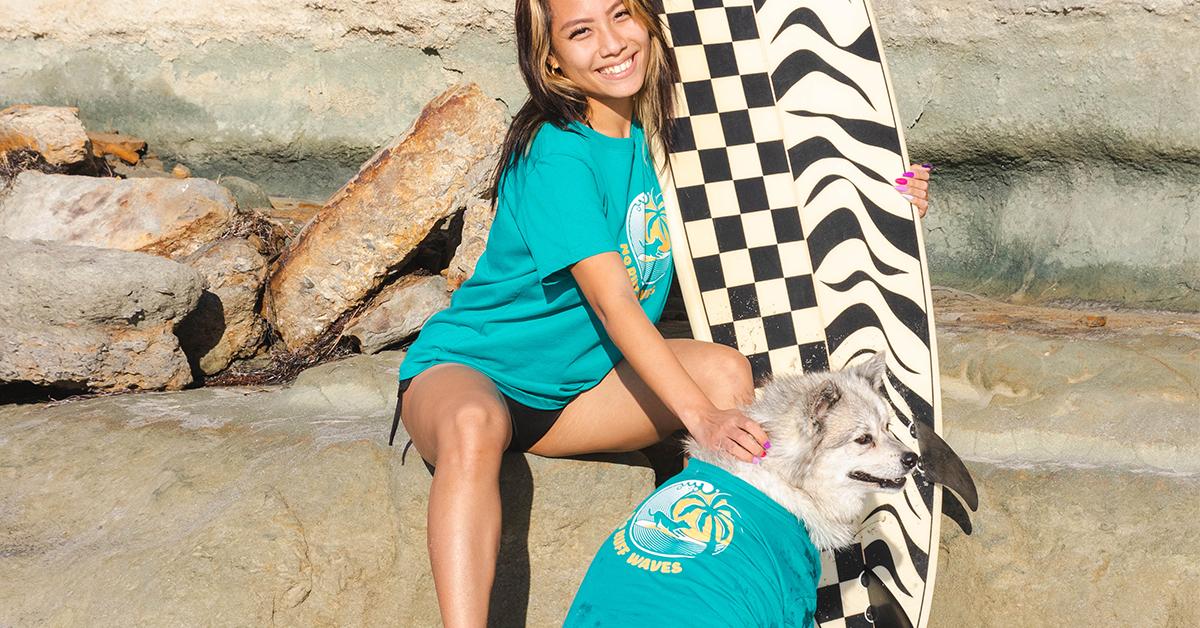 Woman and dog wear teal Fetch The Sun shirts on beach, along with surfboard