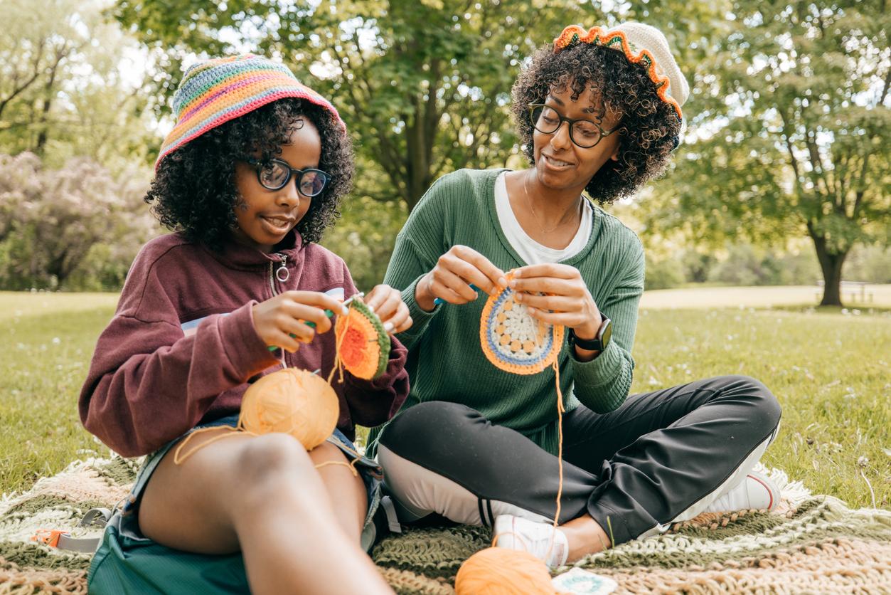 A mother and daughter do crochet and sit on a blanket the grass