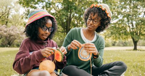 A mother and daughter do crochet and sit on a blanket the grass