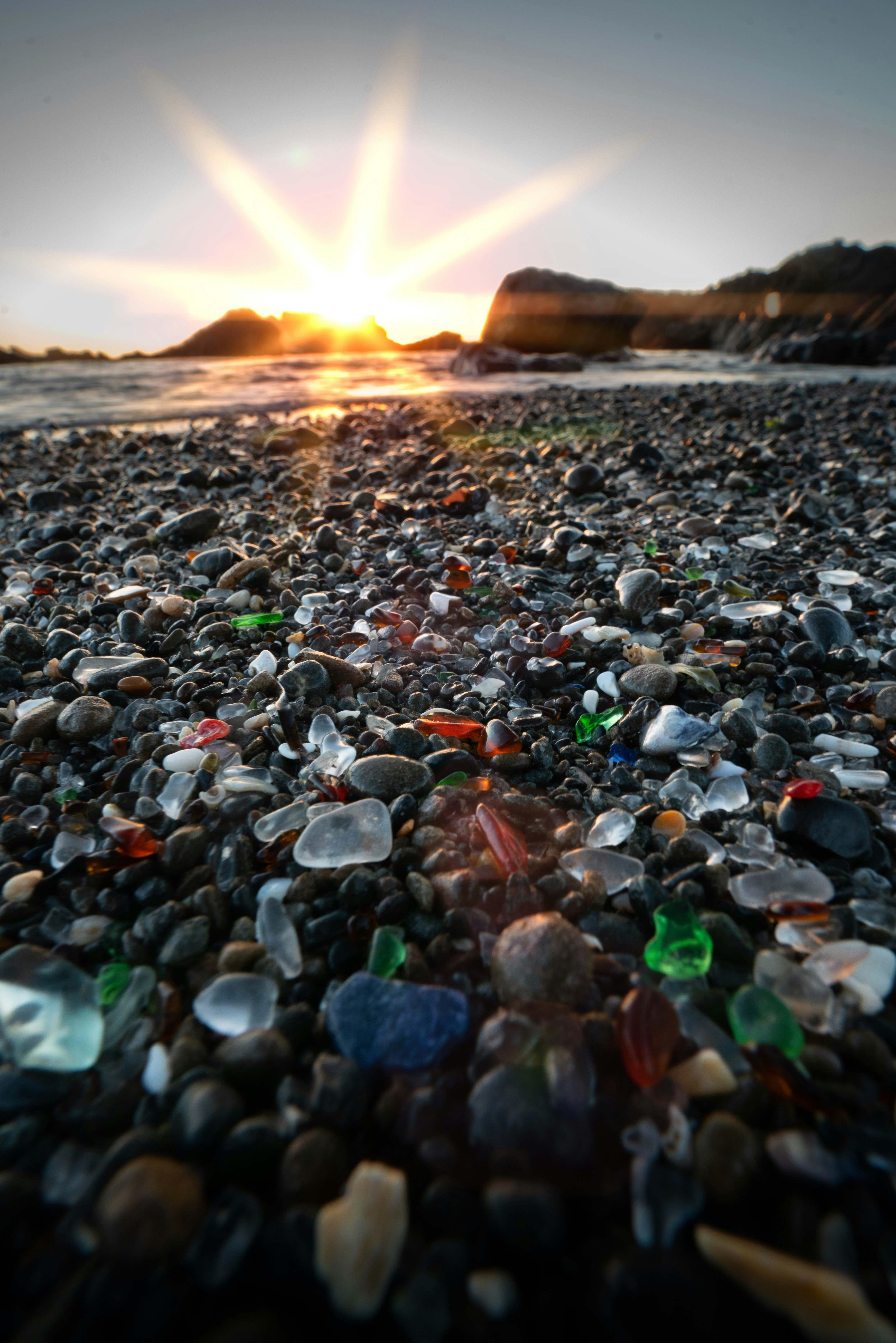 A collection of sea glass is pictured scattered amongst black rocks on a beach with the sunset shining down upon the rocks at the top of the photo.