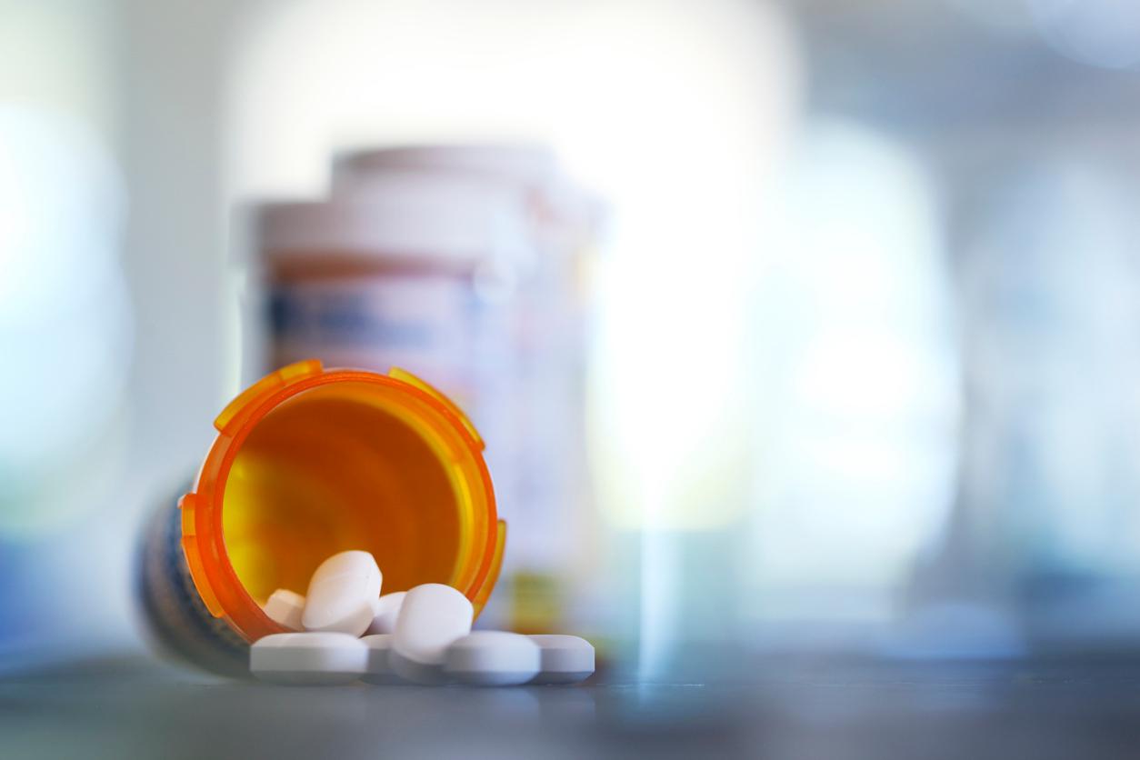 Pills pour out of a prescription bottle onto a counter with other pill bottles in the background.