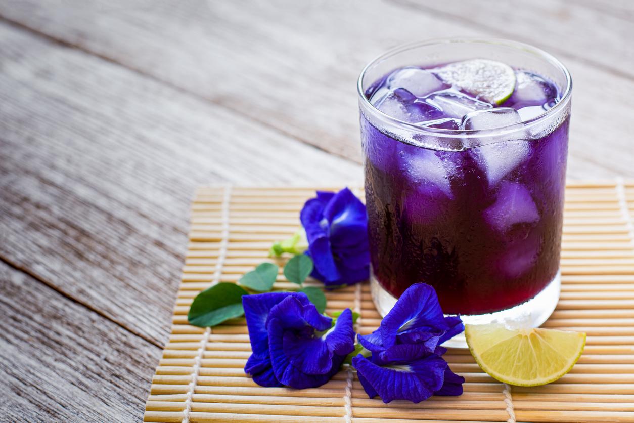 A glass of iced butterfly pea flower tea is pictured with flowers and a lime wedge beside it.