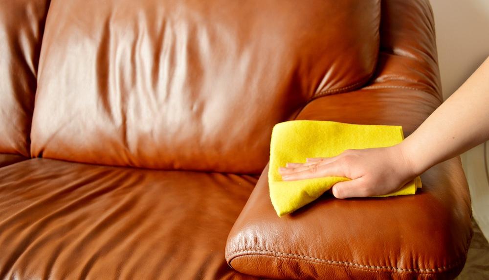 Someone uses a cloth to clean a brown leather couch. 