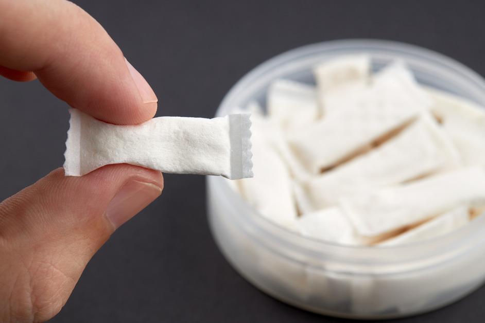 A person holds a nicotine pouch with two fingers with a tin full of the pouches in the background. 