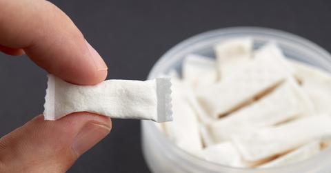 A person holds a nicotine pouch with two fingers with a tin full of the pouches in the background.