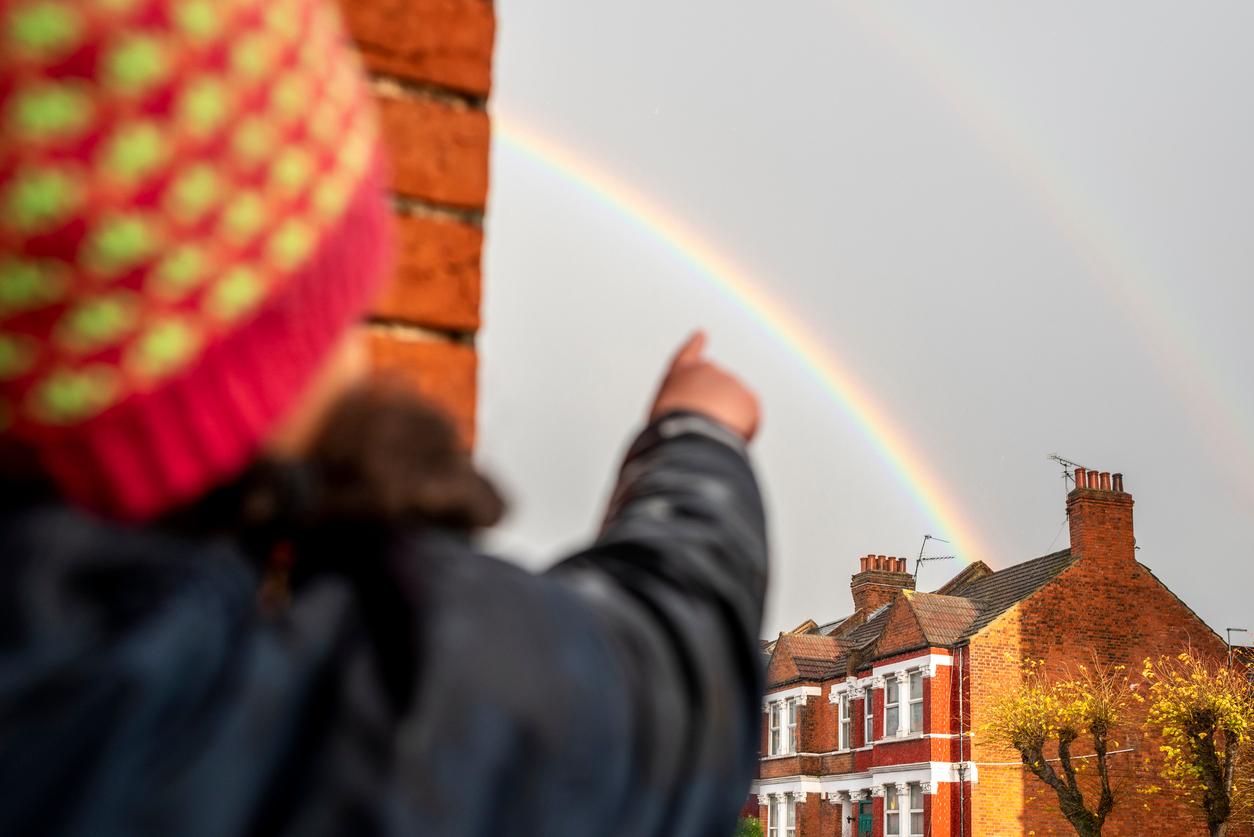 A child in the foreground points to a rainbow over a residential building in the background.