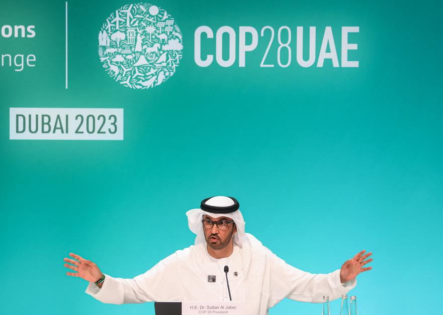 COP28 president Sultan Al Jaber stands at a podium with his hands out wide while talking in front of a screen that says "Dubai 2023."