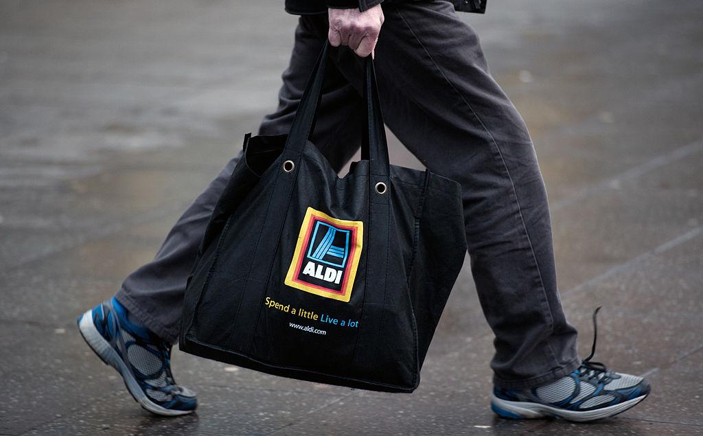 A customer with untied shoelaces and black jeans carries a reusable black Aldi grocery bag in a parking lot.