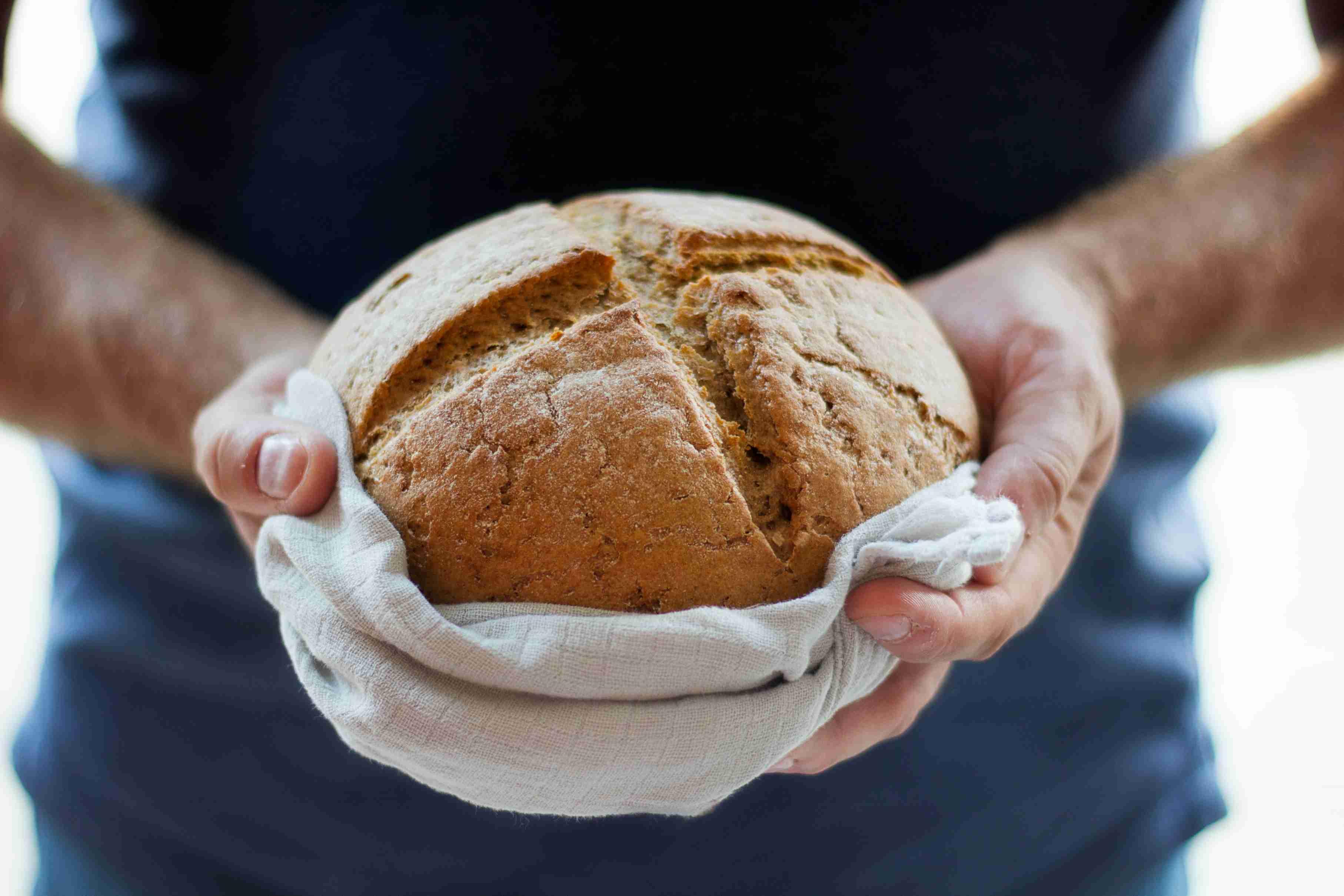 An individual holds out a freshly baked loaf of bread on a towel.