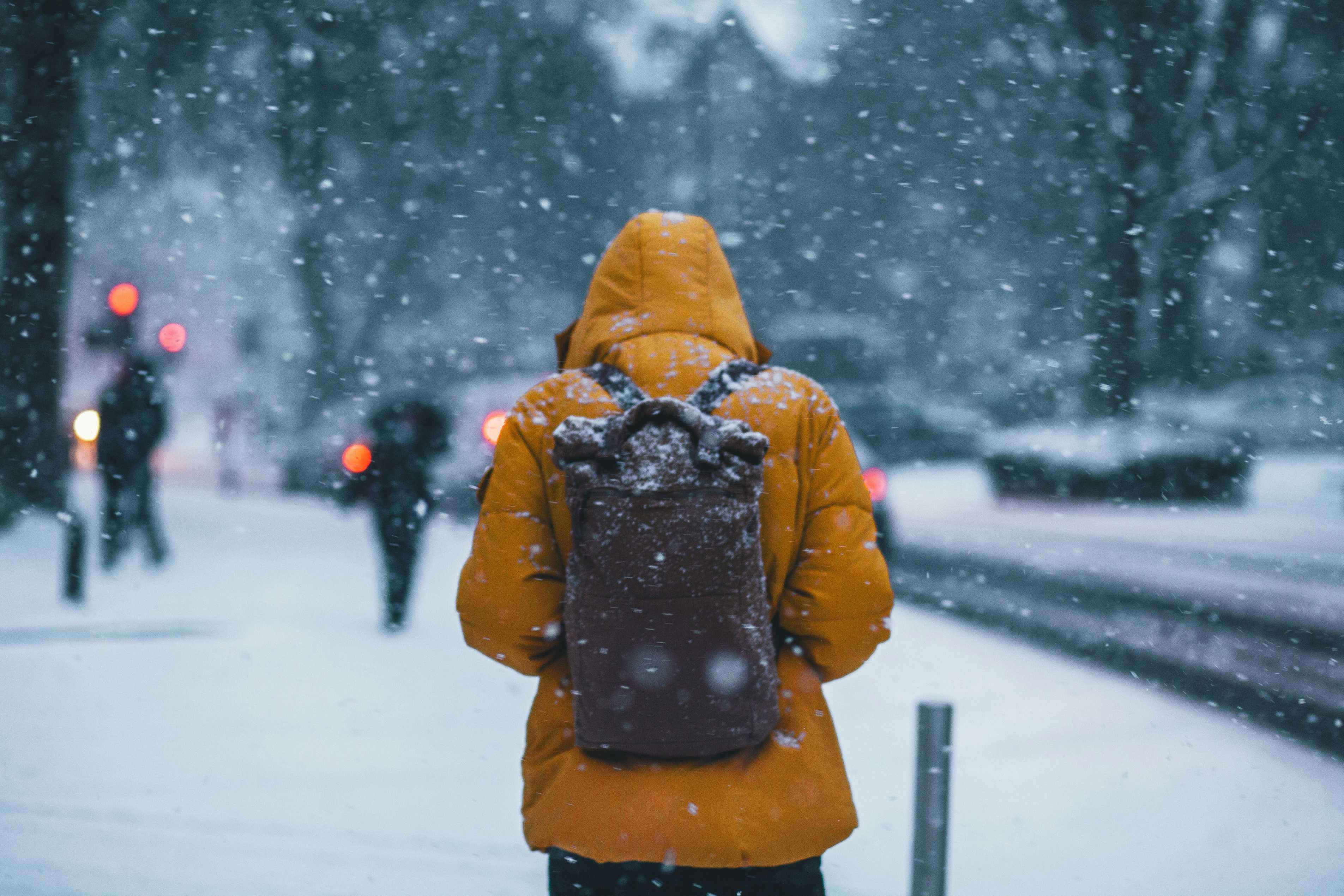 A person in a yellow winter coat and backpack walks through the snow at dusk.