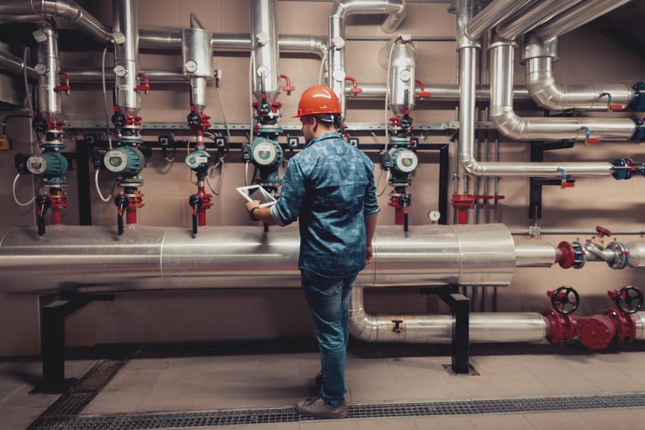 Engineer in orange hat working in a facility with piping on the walls. 