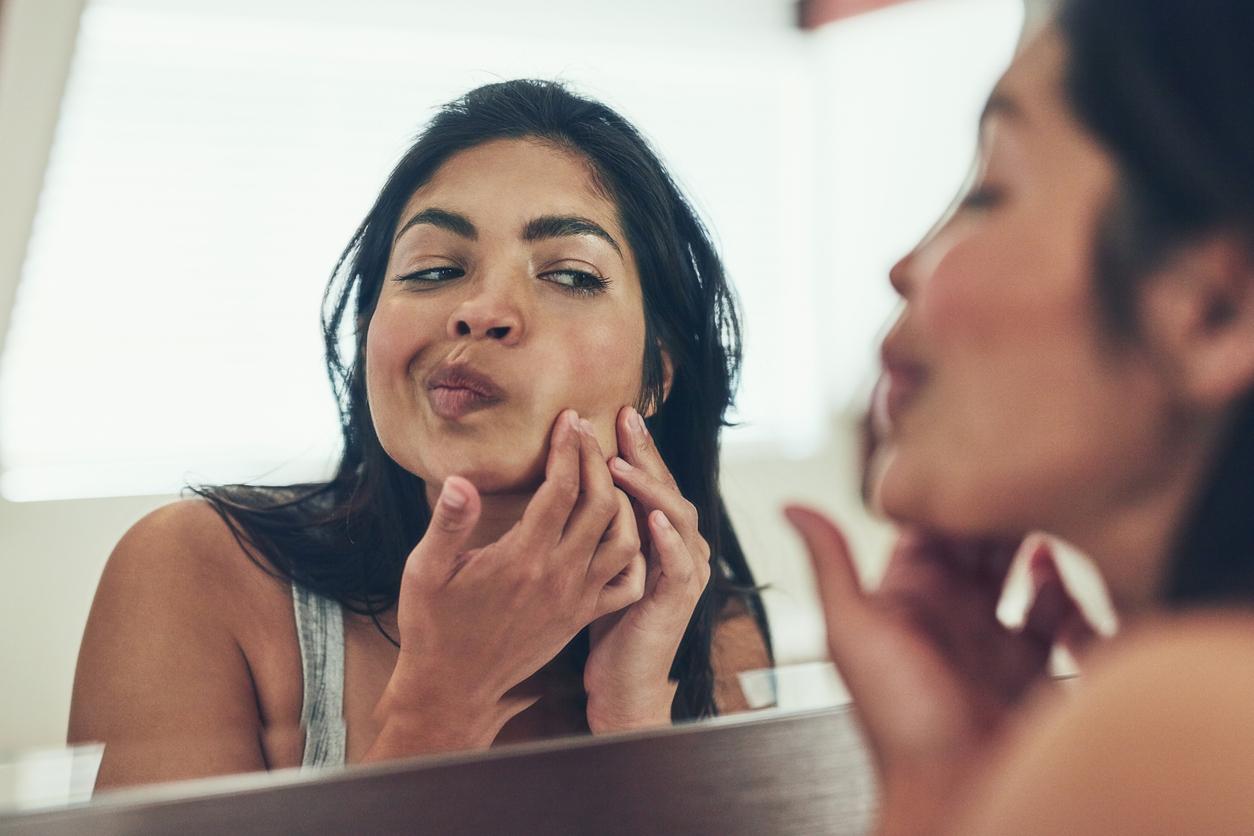 A woman checks a pimple that has appeared on the lower part of her right cheek while looking in the mirror.