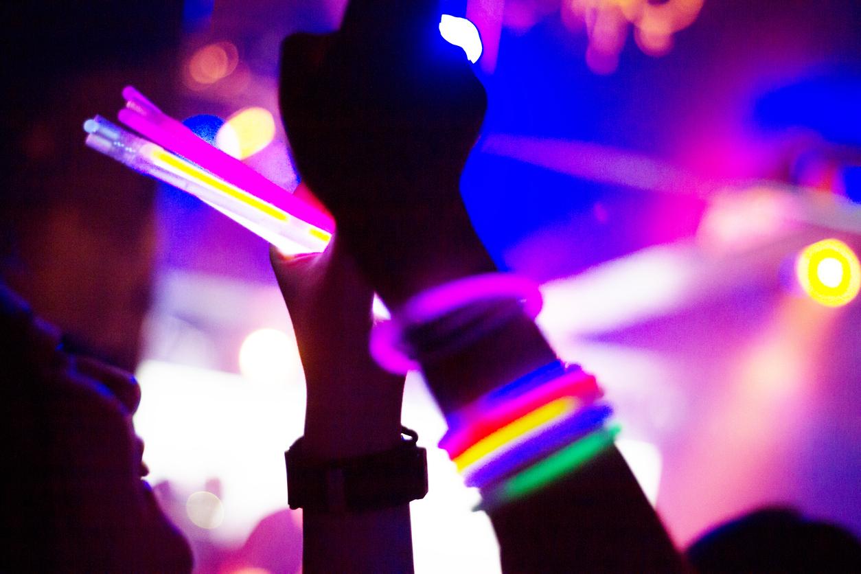 A person at a rave wears multiple glow sticks around their wrist while holding glow sticks in their other hand.