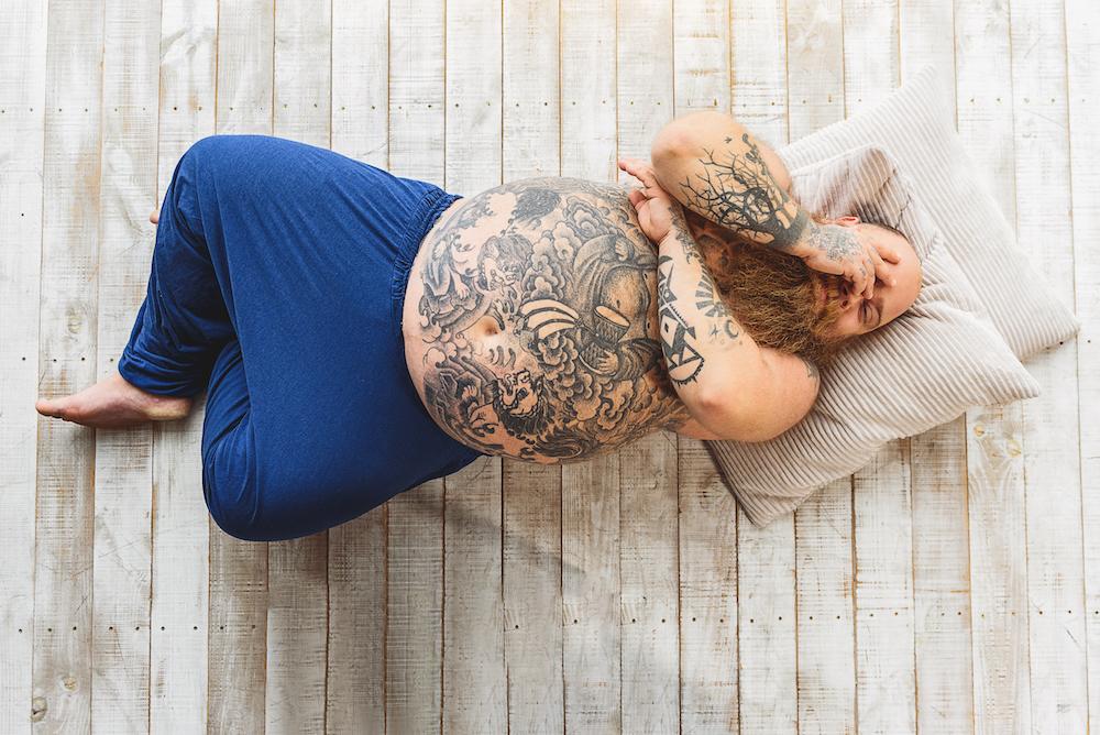 A man with tattoos covering his stomach is laying on the floor covering his face. 