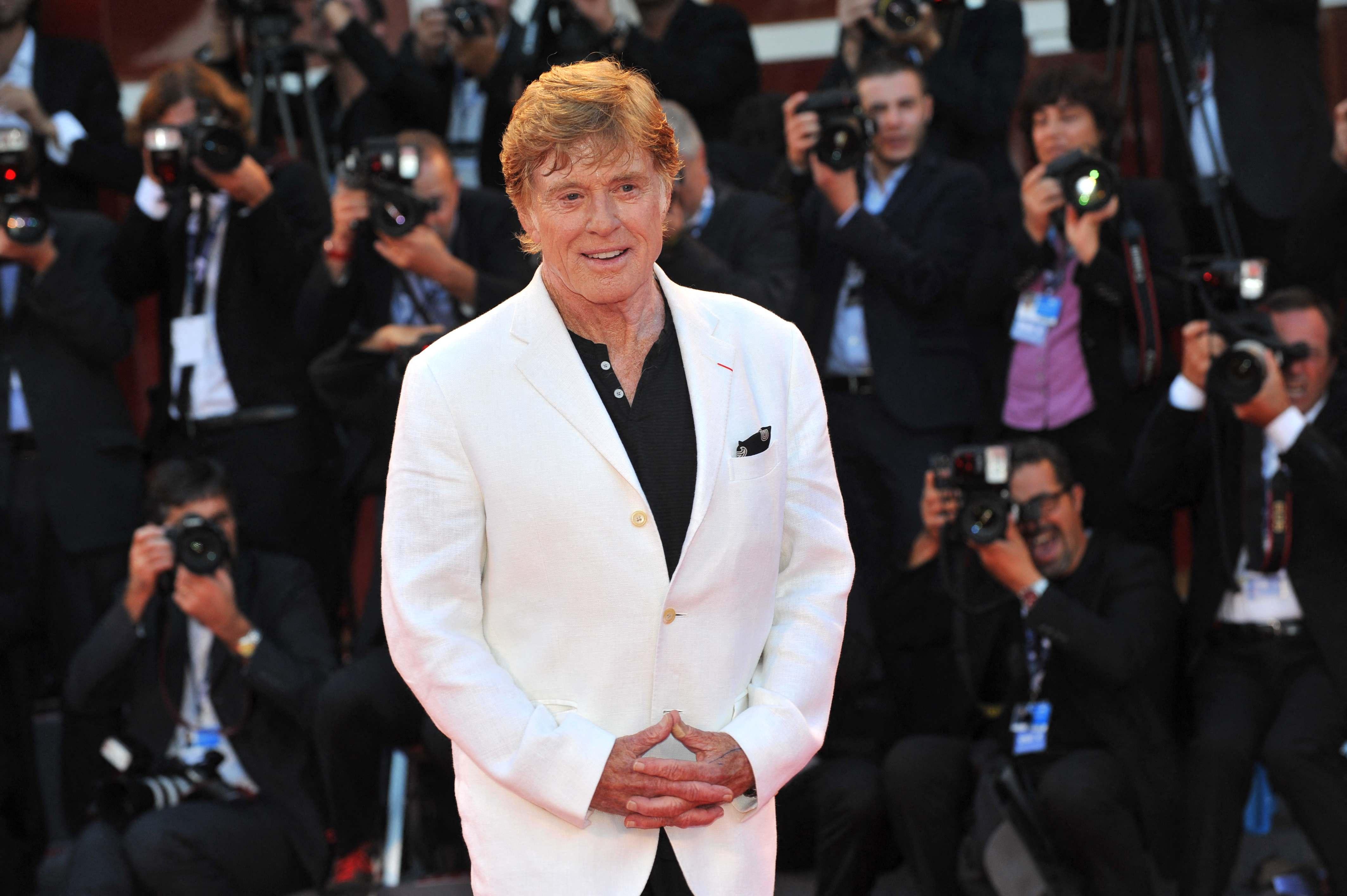 Robert Redford is pictured in a white blazer while standing in front of media members holding cameras.