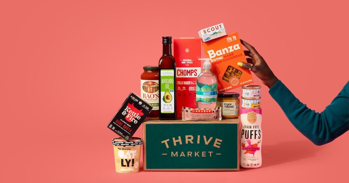 On an orange background, a Thrive Market cardboard box sits with various packaged food products on and next to it, as a hand reaches for a box of Banza pasta.