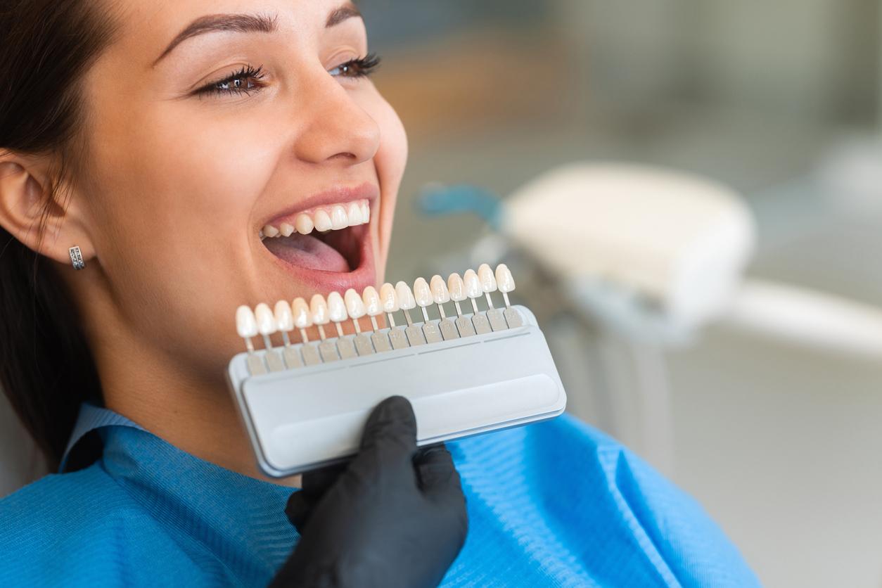 A dentist wearing black gloves uses a palette tooth color sample to match the color of a patient's teeth.