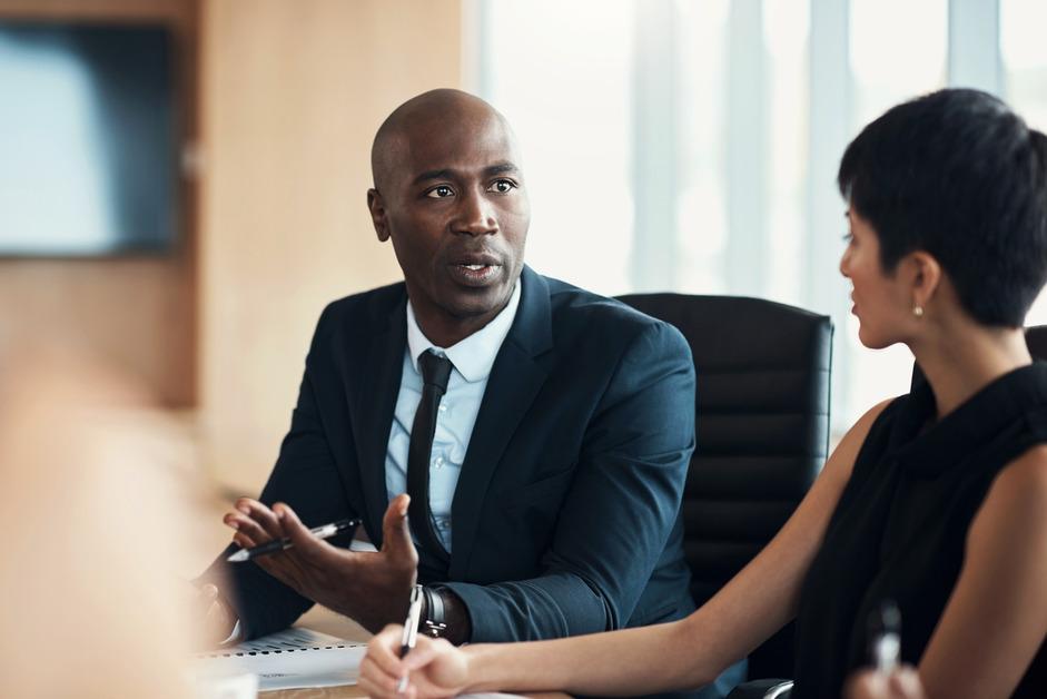 A male lawyer wearing a suit talks to a woman in a conference room while holding a pen. 