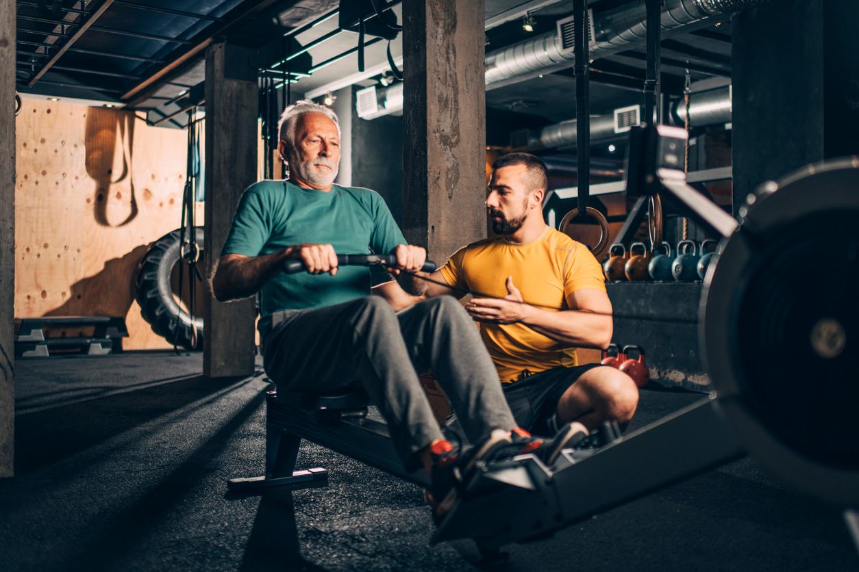 An older adult uses a rowing ergometer while under the supervision of a trainer in a gym.